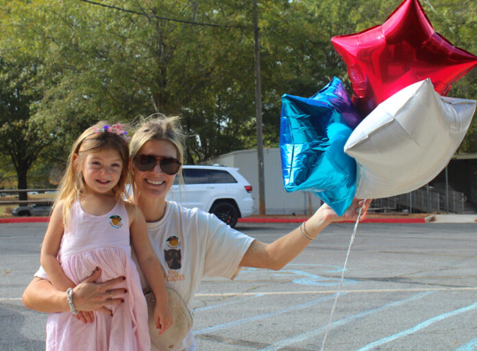 A blonde white woman wearing a white t-shirt and sunglasses kneels on the ground. Her right arm is wrapped around her daughter, a young girl with long hair and a pink dress, and her left hand is holding three balloons, red, white, and blue stars. Both people are wearing Georgia 