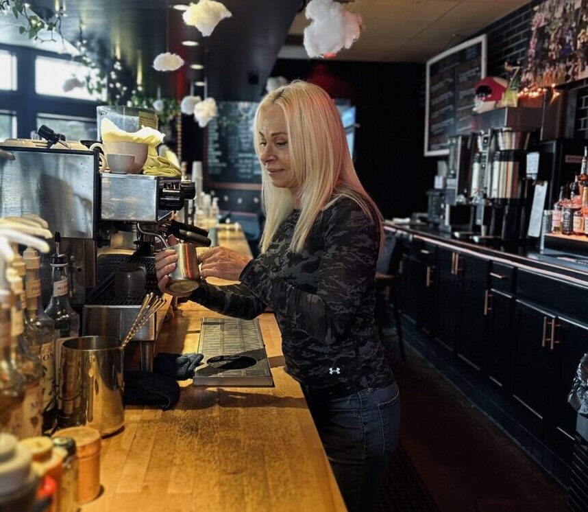 A blonde woman stands at a wooden counter behind a coffee bar making a latte at a coffee machine.