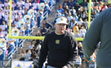 KSU football fans remain angry over head coach firing A white man stands in front of a football goalpost. He is wearing a black long-sleeved shirt with the Kennesaw State University logo. He is wearing a set of headphones and a white baseball hat.