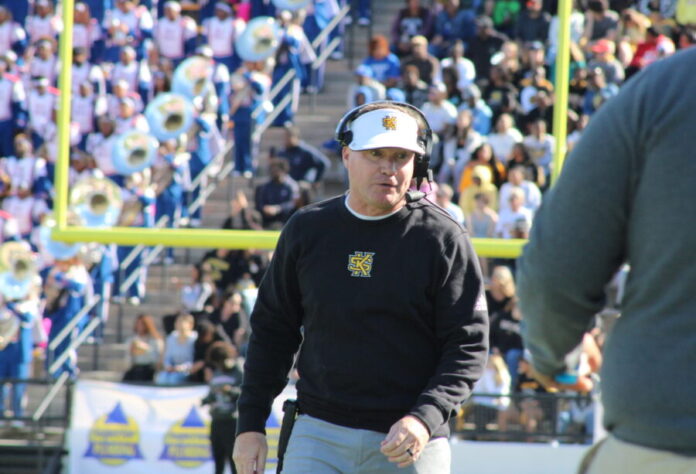 A white man stands in front of a football goalpost. He is wearing a black long-sleeved shirt with the Kennesaw State University logo. He is wearing a set of headphones and a white baseball hat.