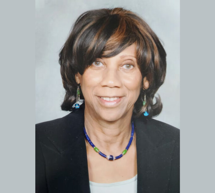 A portrait of a Black woman wearing a black blazer, a blue and green necklace with matching earrings, and straight dark brown hair with highlights cut to her shoulders.