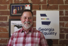 Everyday Hero: Georgia man advocates for LGBTQ health and equality Portrait of a white man with short gray hair and a gray beard. He is wearing a red plaid shirt. He is standing in front of a red brick wall and three framed photos. One has a rainbow outline of Georgia and says "Georgia Equality."