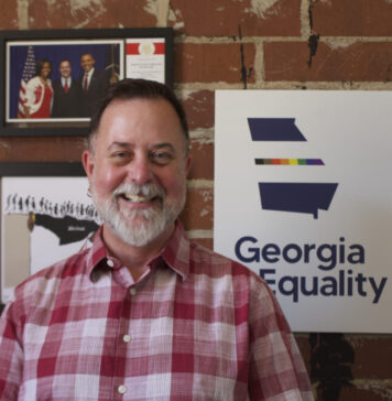 Everyday Hero: Georgia man advocates for LGBTQ health and equality Portrait of a white man with short gray hair and a gray beard. He is wearing a red plaid shirt. He is standing in front of a red brick wall and three framed photos. One has a rainbow outline of Georgia and says "Georgia Equality."