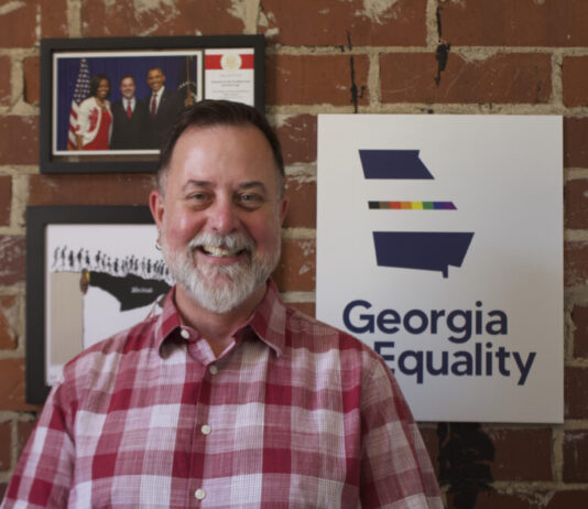 Everyday Hero: Georgia man advocates for LGBTQ health and equality Portrait of a white man with short gray hair and a gray beard. He is wearing a red plaid shirt. He is standing in front of a red brick wall and three framed photos. One has a rainbow outline of Georgia and says "Georgia Equality."