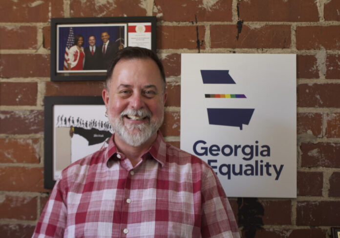 Portrait of a white man with short gray hair and a gray beard. He is wearing a red plaid shirt. He is standing in front of a red brick wall and three framed photos. One has a rainbow outline of Georgia and says 