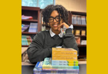 Everyday Hero: Publisher shares stories written by diverse young authors A Black woman with curly hair in an updo smiles at the camera wearing glasses and a grey sweater with a collared shirt underneath. She is sitting at a desk in a library with shelves of books behind her. Her left arm is propped up on a stack of children's books and rests on her face, while her right arm lays on the table.