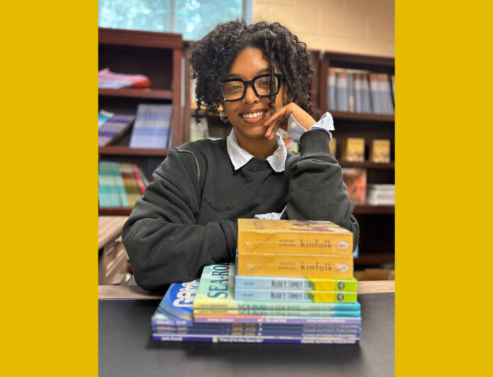 A Black woman with curly hair in an updo smiles at the camera wearing glasses and a grey sweater with a collared shirt underneath. She is sitting at a desk in a library with shelves of books behind her. Her left arm is propped up on a stack of children's books and rests on her face, while her right arm lays on the table.