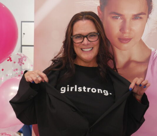 Everyday Hero: Fashion pro empowers next generation of girls A white woman with long wavy dark brown hair stands in front of pink balloons and a photo of a model in pink athletic wear. She is smiling at the camera and wearing a black tee shirt that reads "girlstrong." She is wearing an oversized black blazer and pulling the blazer aside so the text is visible.