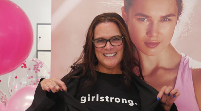 Everyday Hero: Fashion pro empowers next generation of girls A white woman with long wavy dark brown hair stands in front of pink balloons and a photo of a model in pink athletic wear. She is smiling at the camera and wearing a black tee shirt that reads "girlstrong." She is wearing an oversized black blazer and pulling the blazer aside so the text is visible.