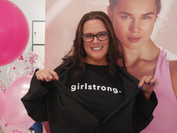 A white woman with long wavy dark brown hair stands in front of pink balloons and a photo of a model in pink athletic wear. She is smiling at the camera and wearing a black tee shirt that reads 