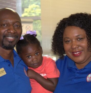 Everyday Hero: Couple uplift students through art, community service An image of three people, shot from the shoulders up, facing the camera and smiling. On the left, an Black adult male with a goatee and braces wears a blue polo and a name tag that reads "Chris A. Woods." In the middle, a young Black girl wearing a pink shirt is held up by Chris. On the right, a Black woman with short hair wears a blue polo.
