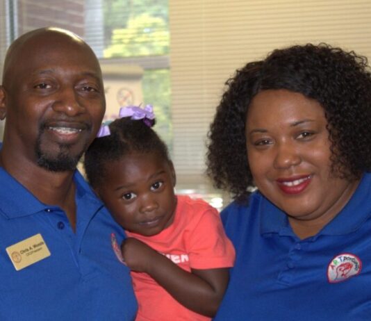 Everyday Hero: Couple uplift students through art, community service An image of three people, shot from the shoulders up, facing the camera and smiling. On the left, an Black adult male with a goatee and braces wears a blue polo and a name tag that reads "Chris A. Woods." In the middle, a young Black girl wearing a pink shirt is held up by Chris. On the right, a Black woman with short hair wears a blue polo.