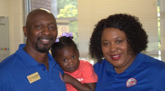 Everyday Hero: Couple uplift students through art, community service An image of three people, shot from the shoulders up, facing the camera and smiling. On the left, an Black adult male with a goatee and braces wears a blue polo and a name tag that reads "Chris A. Woods." In the middle, a young Black girl wearing a pink shirt is held up by Chris. On the right, a Black woman with short hair wears a blue polo.