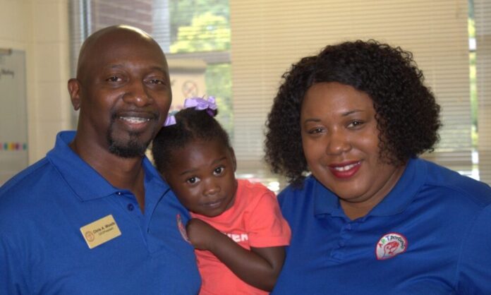 An image of three people, shot from the shoulders up, facing the camera and smiling. On the left, an Black adult male with a goatee and braces wears a blue polo and a name tag that reads 