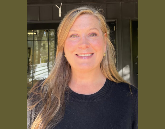 A profile photo of a blonde woman with long hair smiling at the camera. She is wearing a black sweater and small gold earrings. She is standing in front of a dark green building.