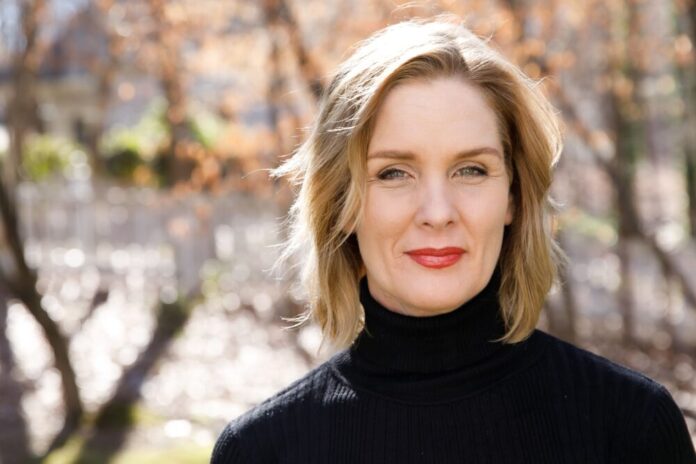 A headshot of a white woman with shoulder-length blonde hair wearing a black turtleneck. She is standing in front of a blurred background of trees.